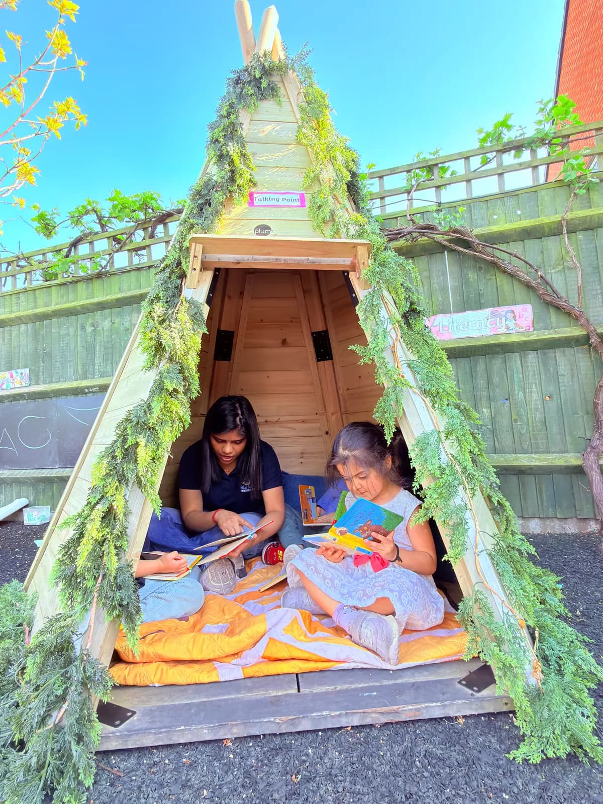 Two children reading inside the leaf-covered teepee in the Blue Nest Montessori Harrow garden